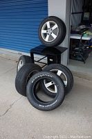 Five tires mounted on alloy rims, one new BF Goodrich Radial T/A tire with label, four used tires showing wear and dirt on rims.