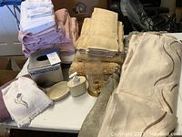 Full view of stacked towels in multiple colors, tissue box cover, soap dish, soap dispenser, decorative glasses, beige rug, and folded shower curtains.