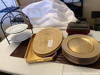 Photo showing all items together including gold chargers stacked, woven placemats, wooden table runner, 2-tier serving stand, gold tray, and covered box.
