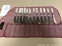 Twelve butter spreaders arranged on burgundy felt roll with one different marked knife at the end and a label card.