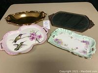 Four decorative trays on a surface. From left to right: gold metal tray, mirrored metal tray with ornate edges, pink hand-painted ceramic tray with hummingbirds, and light green floral ceramic tray.