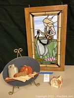 Framed stained glass panel with woman playing string instrument, wooden frame with metal hanging hooks, shown standing behind slate plaque and stone sculpture