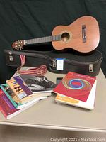 Full lot including Hondo classical guitar, black hard case with latch, guitar strap, and a stack of music books on a table.
