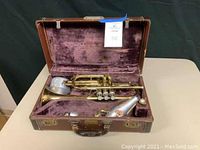 Vintage Conn trumpet stored inside a brown hard case lined with purple velvet. Case lid is open revealing the trumpet and accessories inside.