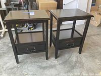 Pair of black painted wooden end tables with drawer and lower shelf on concrete floor in warehouse setting.