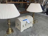 Pair of brass lamps with fluted white fabric shades positioned around a hand-painted decorative trunk, shown on a concrete floor.