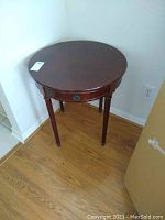 Photo showing full view of round mahogany side table with drawer and four legs on wood floor.