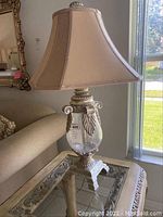 Close-up of one Matter Brothers lamp showing clear glass urn base with decorative acanthus leaf and scroll details, cream fabric shade, and ornate pedestal.