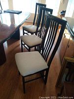 Six black lacquered dining chairs with white upholstered seats arranged around a wooden dining table on hardwood floors. Gloss finish visible with wear.