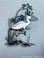 Close-up image of metal and wood patio wall art featuring a white heron bird with detailed features, standing among metalized branches and leaves colored in green and blue.