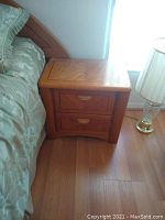 Photo showing one oak nightstand next to a bed and a lamp. Two drawers with wooden integrated handles, medium oak finish.