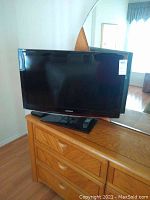 Frontal photo of Samsung 32-inch LCD TV on wooden dresser with remote resting on stand