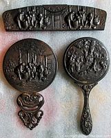 Three piece vanity set laid out flat showing back metal detailing with raised scenes and ornate handles, and amber plastic comb.