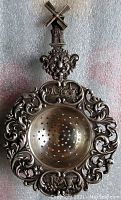 Full view of the silver tea strainer showing ornate floral and landscape designs with a perforated bowl for straining tea and a windmill handle.