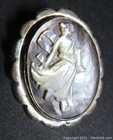 Close-up of front carved mother of pearl cameo brooch showing detailed female figure in flowing robes on iridescent background framed by scalloped 800 silver frame.