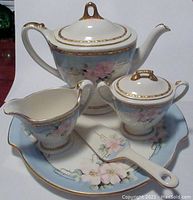 Photo showing the teapot, creamer, sugar bowl, cake plate, and server with floral pattern, blue background, and gold trim.