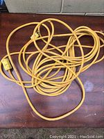 Yellow electrical cord extension placed on a wooden surface, showing the entire length coiled and plug/socket ends visible.