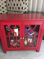 Front view of red wooden cabinet with glass paneled double doors showing geometric shapes and various stored items visible inside.