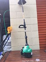 Full view of green gas-powered weed eater with handle against a white wall.
