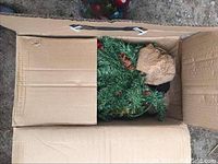 Box containing artificial pine branch wreath with pinecones and burlap bow.