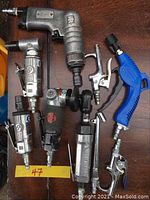 Photo showing seven pneumatic air tools arranged on a wooden surface, including grinders, screwdrivers, and air blowers identified by brand logos and design features.