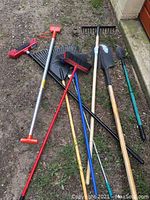 Full view of gardening tools laid on ground, showing nine items including rakes, shovels, picks and brooms