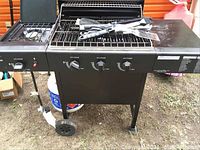 Full front view of black gas grill with three burner control knobs, side burner, wheels, and tripod legs on grass surface.
