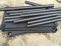 Stack of black cylindrical metal legs with threaded ends arranged on top of several black plastic shelves with rounded ends placed on ground outside.