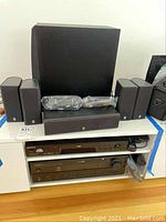 Wide photo showing Yamaha subwoofer, center channel speaker, four small satellite speakers on shelf above Yamaha receiver and Sony DVD player in white shelving unit.