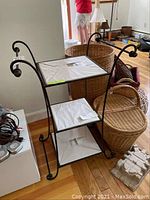 View of the three-tier wrought iron shelf with glass shelves partially covered by paper, set against a wall with various baskets and household items in background.