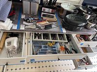 Five drawers showing organized metal flatware, assorted kitchen utensils (ladles, peelers, tongs), cutting boards, knives block, spice grinder and pots and pans on the countertop.