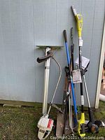 Photo of Ryobi tree saw and multiple gardening tools including shovels, rakes, and trimmers leaning against a shed.