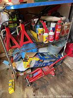 Metal shelf loaded with various automotive tools, fluids, and red metal jack stands visible, hydraulic floor jack on floor below shelf
