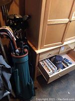 Two golf bags are shown beside a wooden cabinet and other items. The front green bag holds multiple golf clubs including a club with Cleveland Launch 905 headcover. Behind it is a black golf bag.