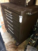 Front and side view showing nine drawers with silver horizontal handles, brown metal surface with some scratches and wear, mounted on wheels.
