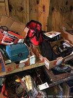 Overview of power tools stored in cardboard boxes and tool bags including Black & Decker and Makita tools.