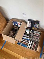 Box containing mixed CDs and DVDs arranged in rows. Visible CDs and DVDs clearly show variety of artists and titles.