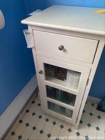 Front view of white bathroom cabinet showing one drawer and cupboard door with three textured glass panels. Cabinet has metal knobs and sits on tiled floor.