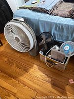 Five assorted electric fans sitting on wooden floor beside a bed with a blue bedspread, including a large Lasko Cyclone box fan with remote on top, a small white clip fan, and three others in various shapes and sizes.