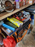 Box filled with orange extension cords, gray hanging work lights, and assorted electrical wires on a shelf above a General generator