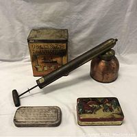 Photo showing four antique items: brass spray can with long nozzle and pump handle, Ocean Blend Indian Head Tea tin can, decorative tin can with winter scene, and metal straight razor case.
