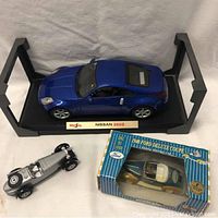 Blue Maisto Nissan 350Z model car on display base with nameplate, alongside other toy cars and boxed collectible in background.