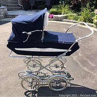 Side view of antique baby carriage showing navy blue fabric, white interior lining, collapsible hood, chrome frame, and large spoked wheels.