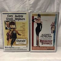 Two vintage movie posters side by side in hard plastic sleeves, one for 'Charade' and one for 'Breakfast at Tiffany's'.