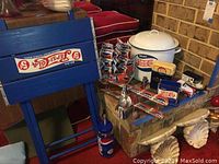 Photo showing a blue wooden crate with vintage Pepsi branding, multiple Pepsi cans, toy trucks, and the white enamel drink dispenser placed on a brick hearth.