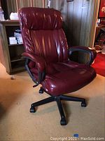 Full view of maroon padded office chair with black base placed on carpeted floor in front of wooden shelving.