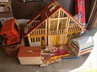 Wide shot showing large wooden Barbie dollhouse with red roof, orange vehicle toy, white and blue friend ship camper playset, and a wooden box beside dollhouse with scattered parts.
