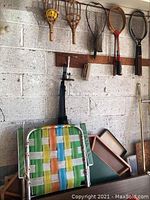 Photo of vintage wood-framed rackets and nets hung on a wall above a green and yellow woven folding lawn chair.