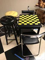 Photo showing yellow and black padded tufted barstools and checkerboard painted game table with two folding chairs.