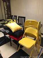 Photo showing four yellow metal folding chairs stacked together with visible signs of wear. Two black metal back chairs with red padded seats visible behind holding various throw pillows and a folded afghan on top.